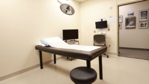 Medical examination room equipped with a brown examination table, bright overhead light, TV, chair, and wall-mounted equipment.