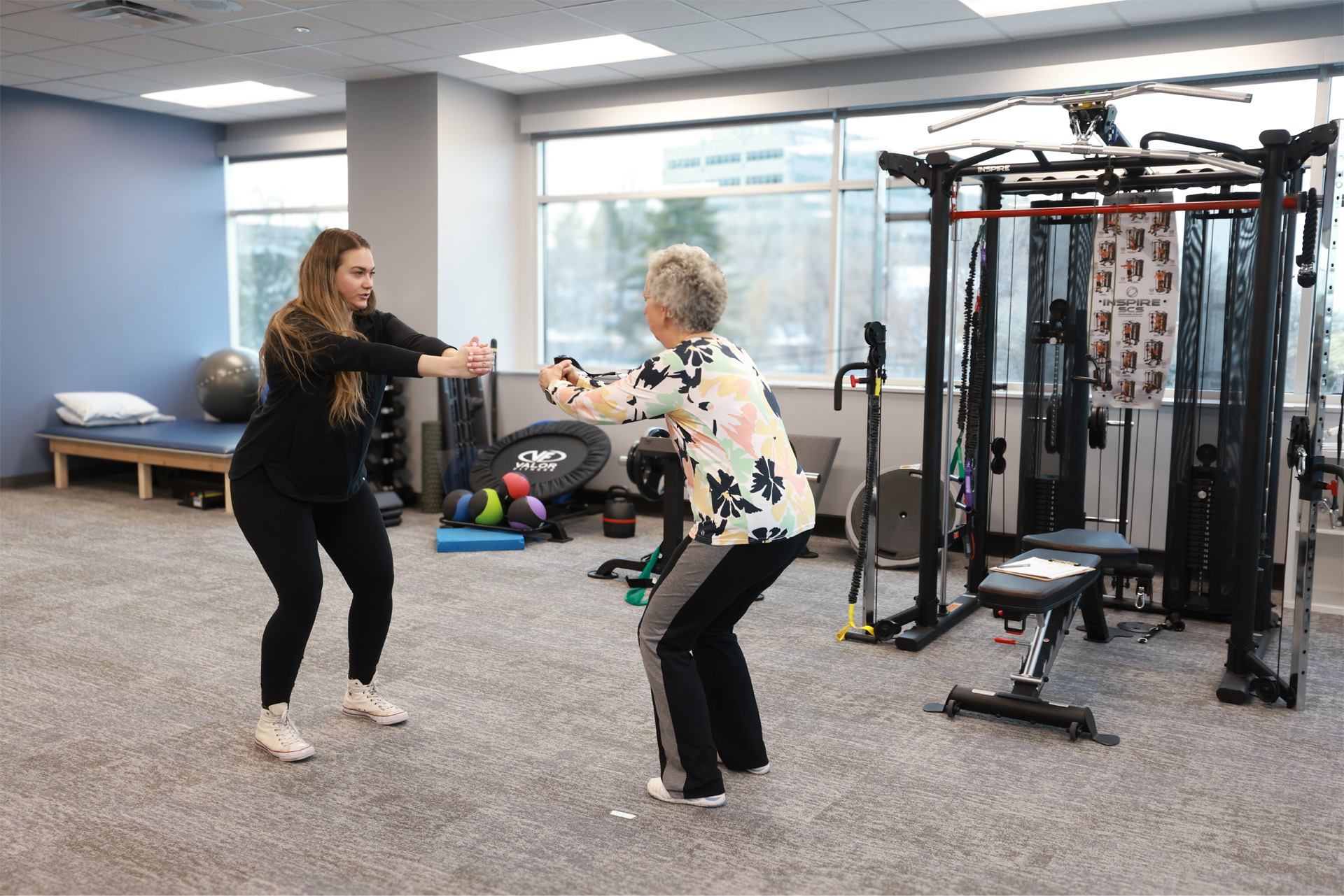 A physical therapist and an elderly patient performing squats together inside a gym equipped with weight machine, weighted balls and other exercise equipment.