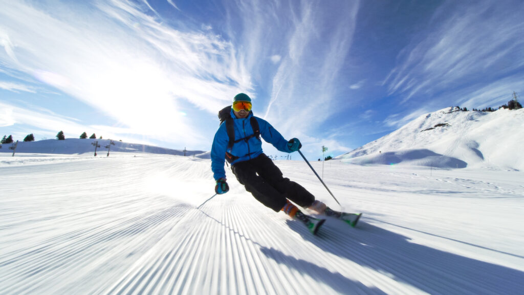 A male skier in a blue jacket and black pants glides down a sunlit, groomed slope, surrounded by snowy mountains and a clear blue sky.
