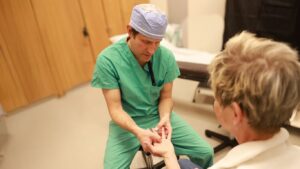A doctor wearing green scrubs and medical cap examining an elderly patient's hand and fingers.