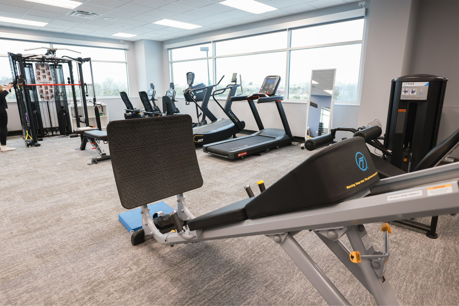 A bright gym room with large windows providing natural light equipped with modern exercise machine including treadmills, a weight machine, and a rowing machine.