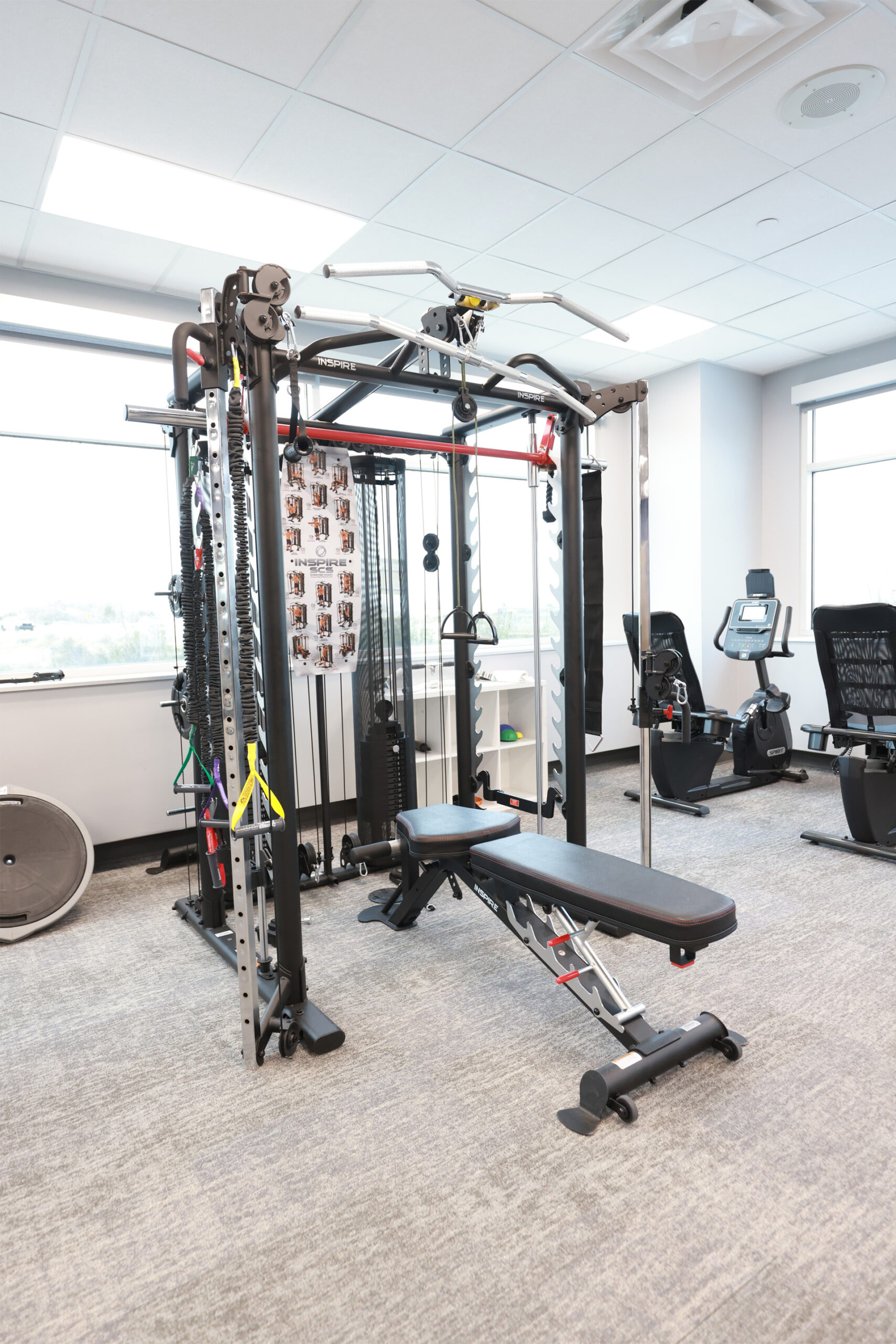 Bright, spacious and modern gym with large windows equipped with weight machine, adjustable bench, and exercise bike.